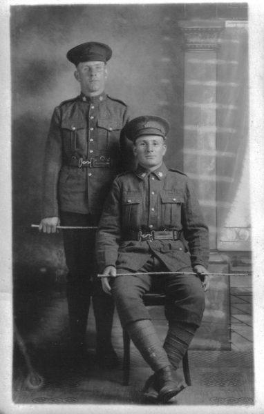 Two soldiers in uniform, one seated, the other standing, in a vintage photograph.