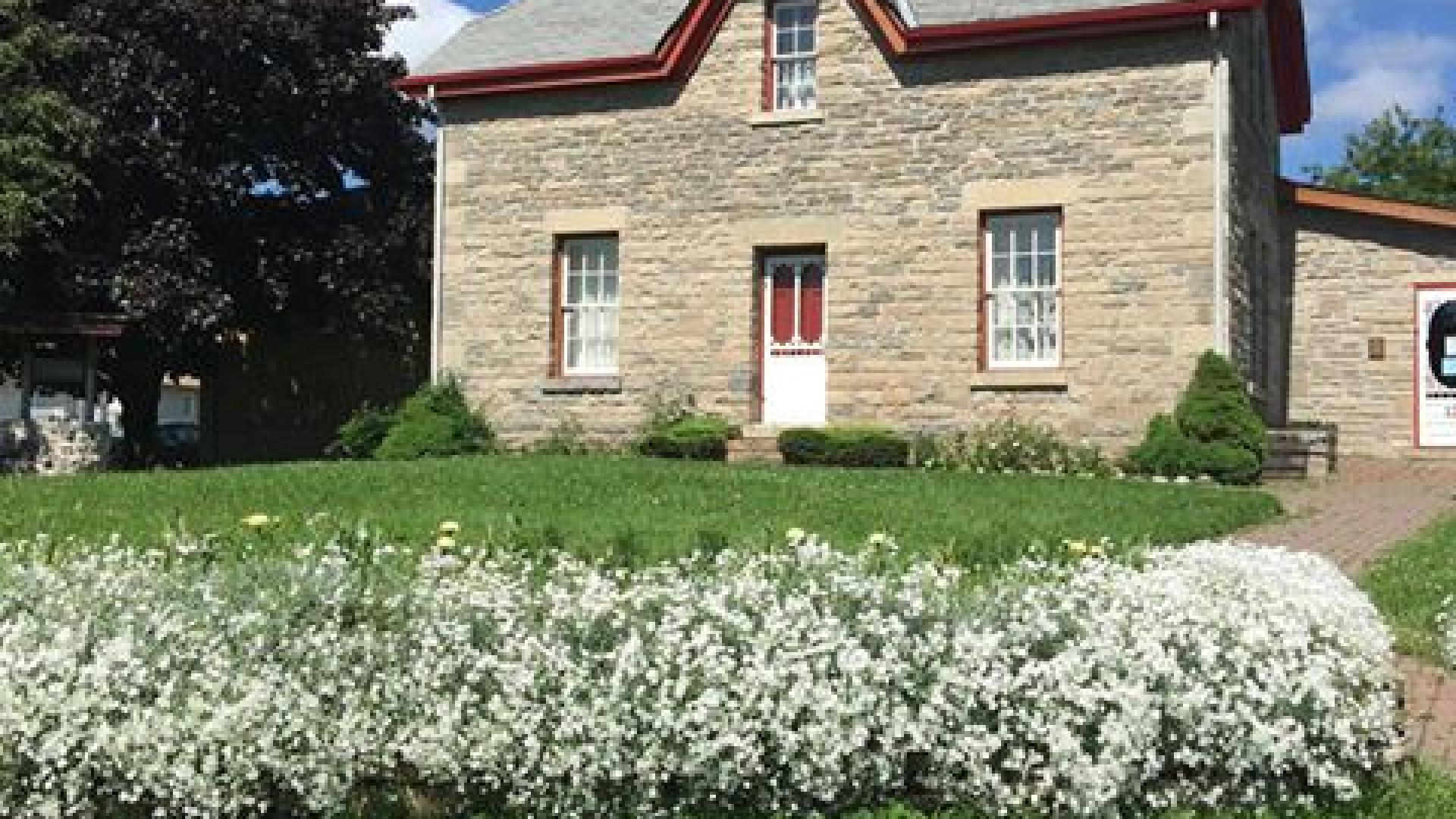 Assiginack musem with red trim, lush garden, under a blue sky with clouds.