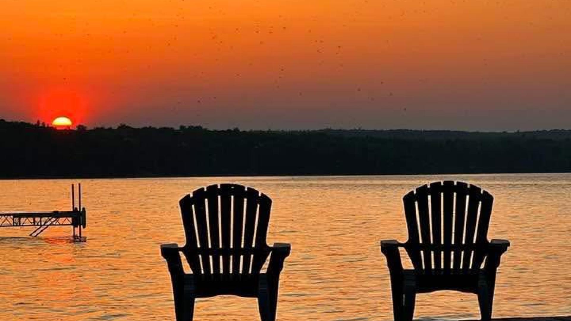 two Muskoka style chairs side by side on a dock with a sun rising over the bay