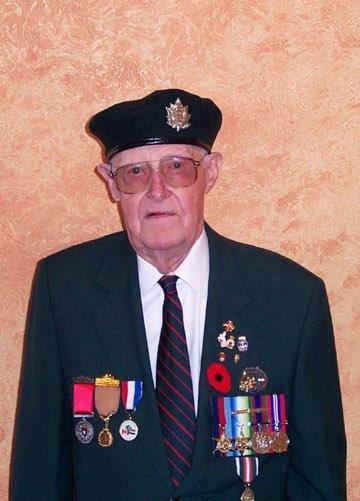 Elderly man in uniform with medals, wearing a beret, standing against a textured wall.