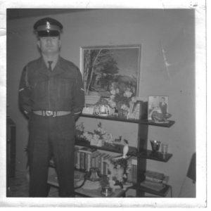 Man in uniform stands in front of a bookshelf in a living room, black and white photograph.
