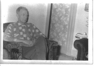 Man sitting in an armchair, wearing a checkered sweater, beside floral wallpaper. Black and white photo.