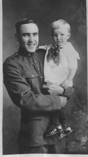 Man in uniform smiling, holding a young child wearing a dress and boots. Black-and-white photo.