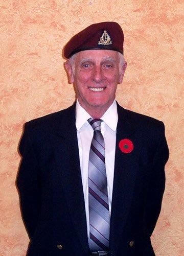 Elderly man in suit and beret with red poppy on lapel, smiling against a textured wall.