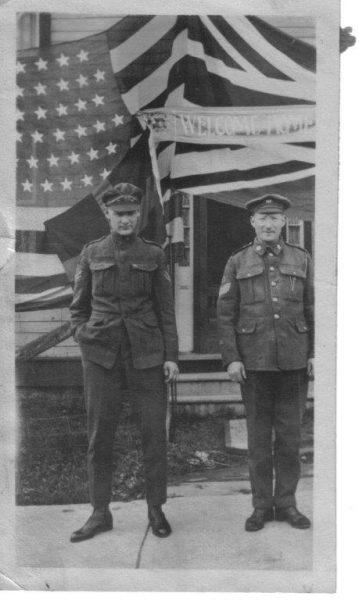 Two soldiers in uniform stand in front of a large American flag.