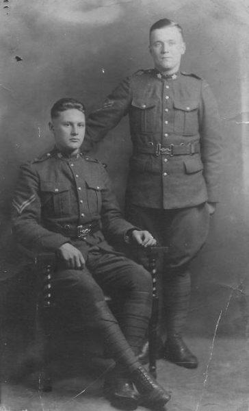 Two soldiers in vintage military uniforms, one seated, one standing. Black and white photograph.