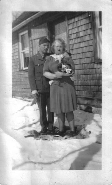 Couple in winter clothes holding a dog, standing on snow by a wooden house.