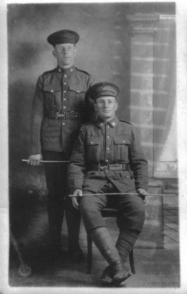 Two soldiers in uniform, one seated, one standing, both with hats, sepia-toned photo.