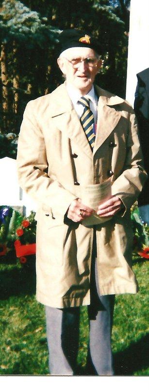 Elderly man in a tan trench coat and striped tie, standing outdoors.