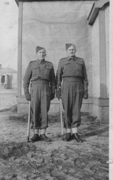 Two soldiers in uniform stand on crutches outside a building.