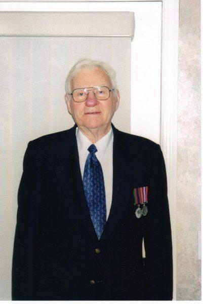 Elderly man in a suit with medals, standing indoors, smiling slightly.