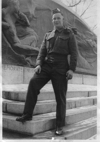 Soldier standing on steps, wearing uniform and hat, in front of war memorial sculpture.