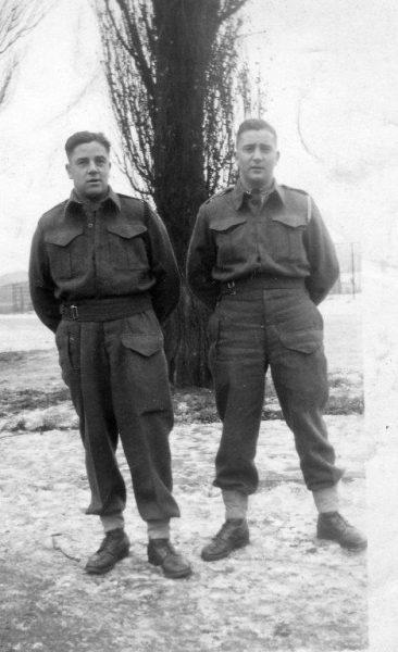 Two soldiers in uniforms stand outside on a snowy day, tree in the background, black and white photo.