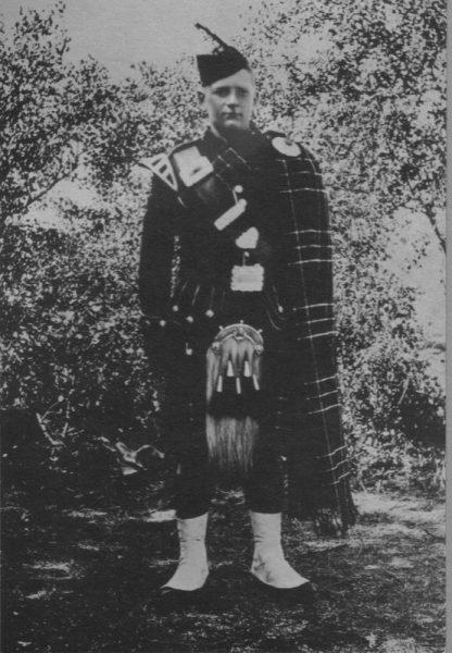 Scottish Highlander in traditional attire stands outdoors.