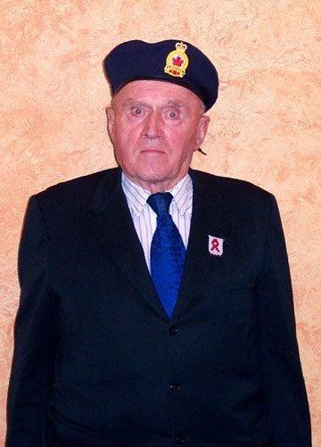 Elderly man in suit and beret, standing against a beige wall.