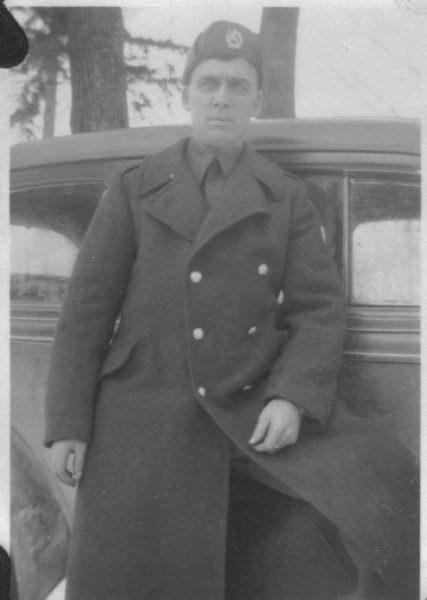 Man in military coat leans against a vintage car, trees in background. Black and white photo.