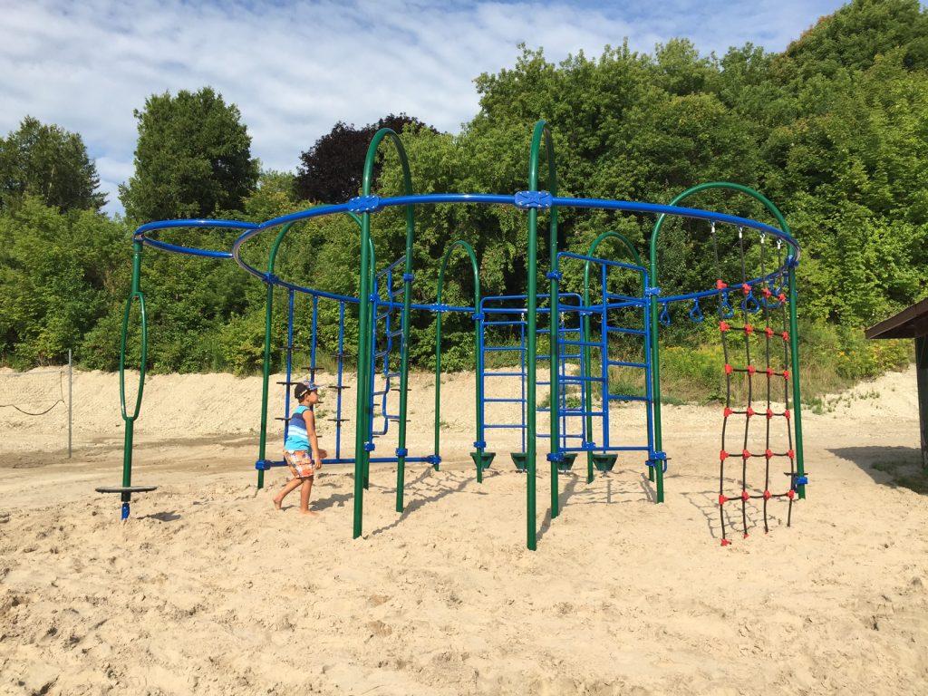 Playground climbing structure on sandy ground, surrounded by trees.