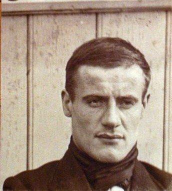 Sepia-tone portrait of a man with short hair, wearing a dark jacket, standing against wooden panels.