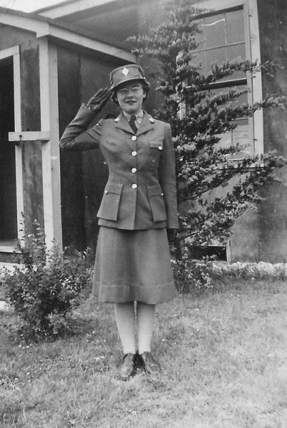 A woman in military uniform saluting, standing in front of a building.