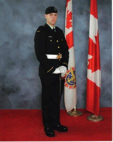Military officer in dress uniform stands beside Canadian flags.