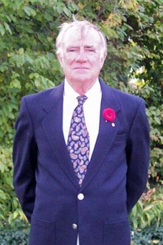Elderly man in a suit with a red poppy, standing against greenery.