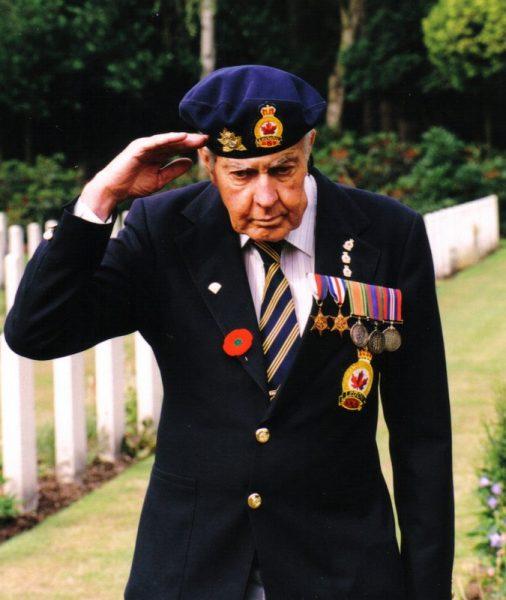 Elderly man in uniform saluting, wearing medals and a beret, in a cemetery.