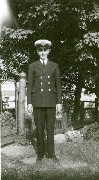 Young man in naval uniform standing in a garden, smiling.