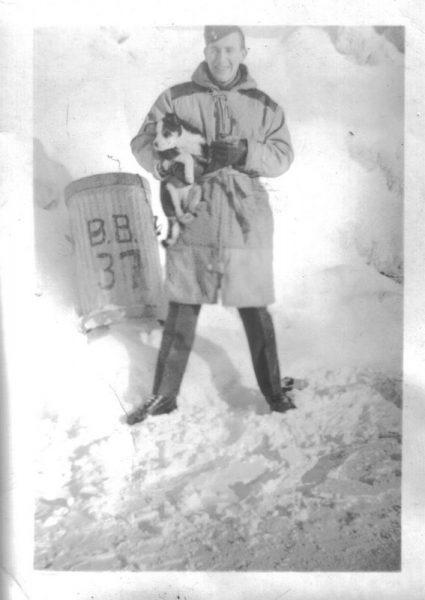 Man in winter coat holding a dog, standing in the snow near a marked pole.