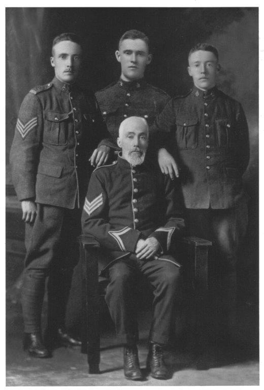 Four men in vintage military uniforms, one seated, three standing behind. Black and white photo.